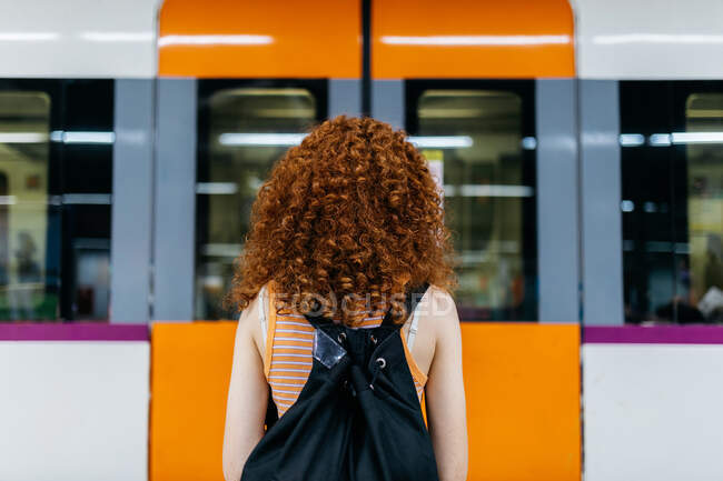 Vista trasera de turista femenina sin rostro con mochila y pelo rizado contra tren subterráneo en terminal - foto de stock