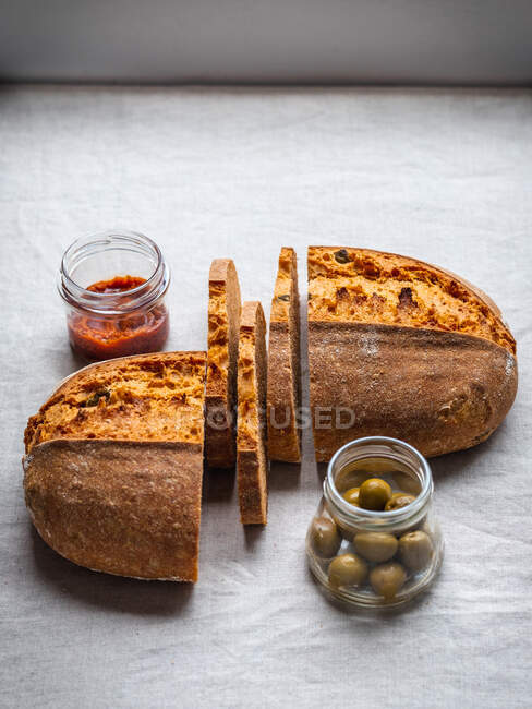 From above cut loaf of bread placed on table near red pesto sauce and jar of green olives — Stock Photo
