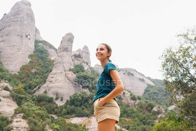 Vista laterale di allegra viaggiatrice con le mani sui fianchi che contempla Montserrat con gli alberi mentre distoglie lo sguardo durante un'escursione in Spagna — Foto stock