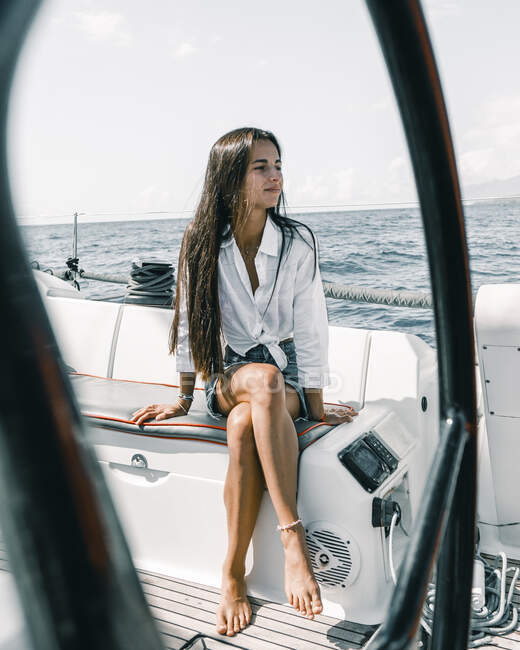 Contemplative female teenager sitting with crossed legs on bench of motorboat on ocean while looking away in Tenerife Spain — Stock Photo
