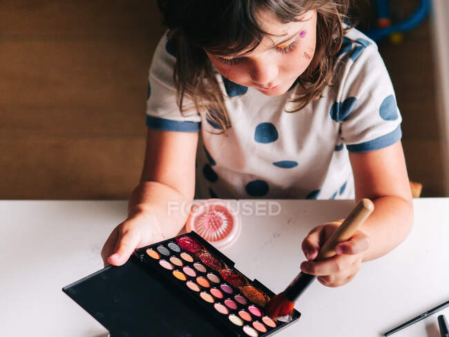 From above kid with applicator making up face at table with assorted cosmetic products in house — Stock Photo