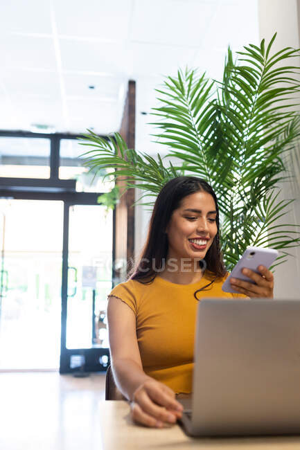 Happy female freelancer sitting at wooden table in cafe browsing non mobile phone while typing on netbook — Stock Photo