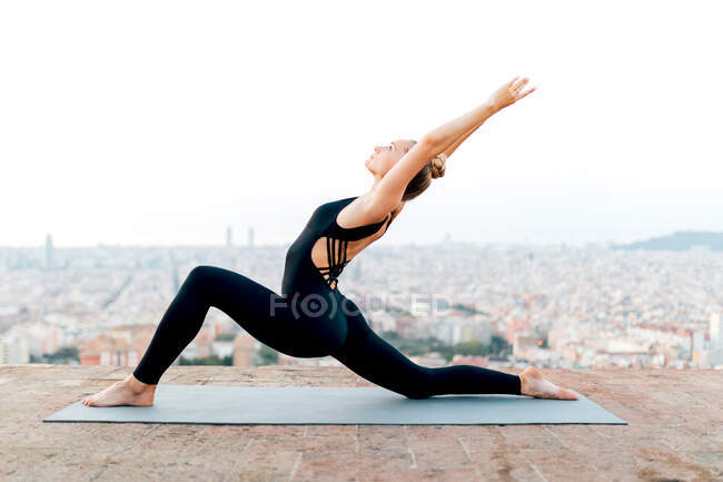 Вид збоку на повне тіло босоніж мирної жінки з піднятими руками, що практикують Півмісяць Лаунж на коліні з лабіринтом Вгору на даху — стокове фото