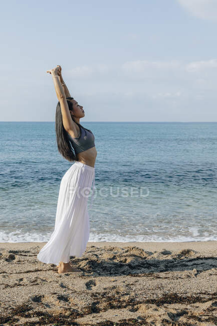 Vista lateral da jovem etnia feminina rachando juntas durante a prática de ioga na praia de areia contra o oceano à luz do sol — Fotografia de Stock