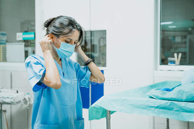 Mujer veterinaria en uniforme que se pone mascarilla desechable mientras mira hacia abajo en el trabajo en la clínica - foto de stock