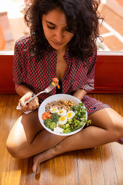Alto ángulo de hembra joven con el pelo rizado sentado con las piernas cruzadas y comer super plato de comida con garbanzo huevos picados brócoli - foto de stock