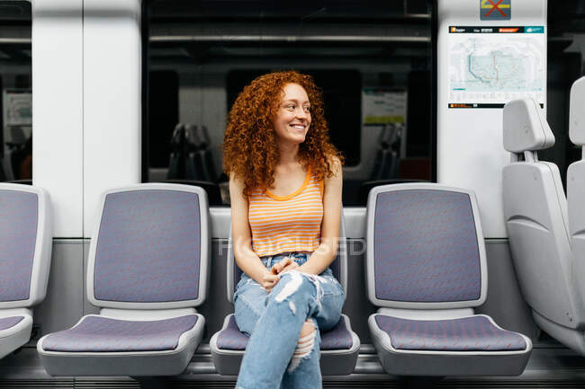 Mujer joven contenta en jeans rasgados con el pelo rojo rizado mirando hacia otro lado en el asiento mientras viaja en tren - foto de stock