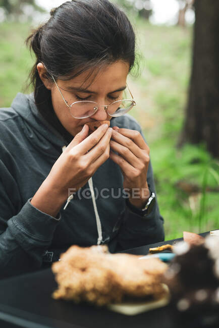 Femme calme aux lunettes assise à table et reniflant des champignons frais dans la forêt pendant la journée — Photo de stock
