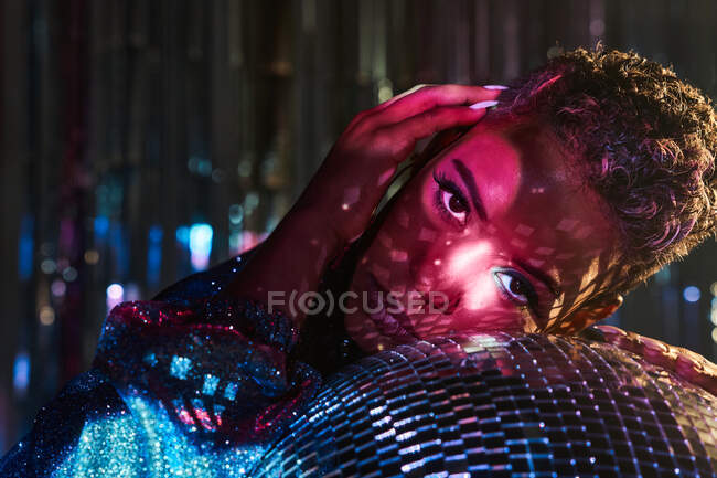 Trendy young ethnic female touching face while looking at camera and leaning on mirror ball in nightclub — Stock Photo