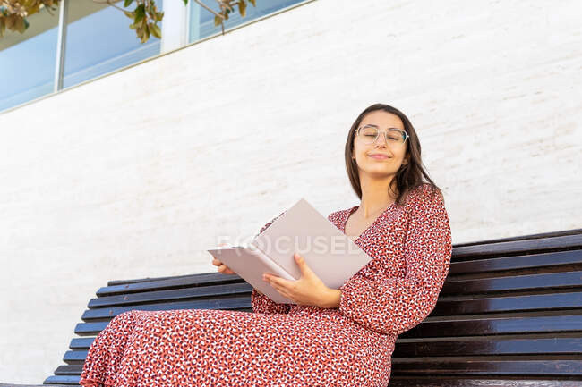 Positive junge Frau in stilvoller Kleidung sitzt mit aufgeschlagenem Buch auf Holzbank gegen Gebäude mit Lichtwand bei Tag mit geschlossenen Augen — Stockfoto