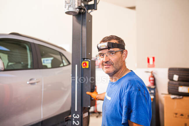 Side view of cheerful mature male technician operating elevator with modern automobile in repair service and looking at camera — Stock Photo