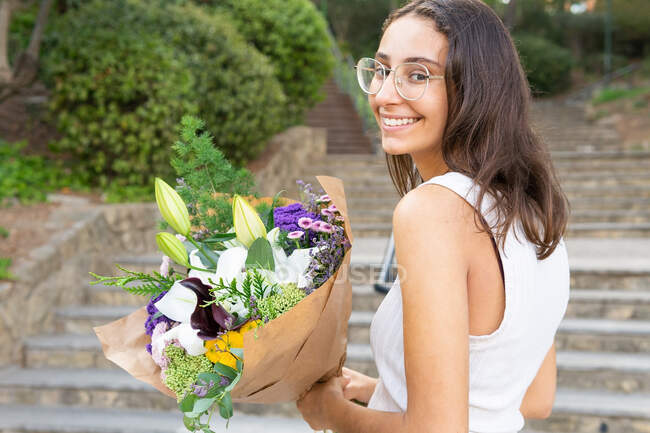 Contenuto giovane femmina in occhiali guardando la macchina fotografica in piedi con mazzo di fiori in fiore sulle scale urbane — Foto stock