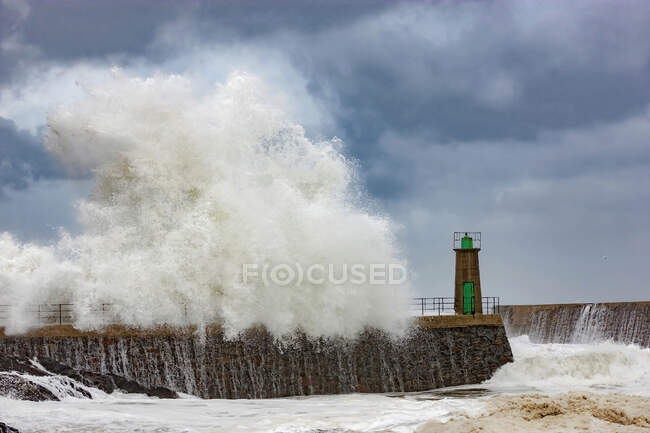 Huge foamy sea waves crashing against stony breakwater with old lighthouse tower against blue cloudy sky in Port of Viavelez in Asturias Spain — Stock Photo