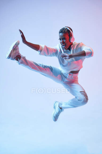 Mujer étnica enérgica en auriculares inalámbricos y ropa de moda saltando con la pierna levantada y la boca abierta mientras baila hip hop - foto de stock