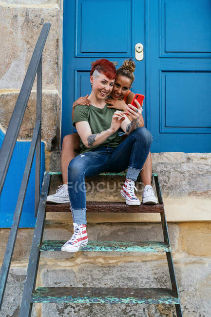 Jovens mulheres homossexuais alegres em roupas da moda navegando na internet no celular enquanto abraçam na escada contra a casa — Fotografia de Stock
