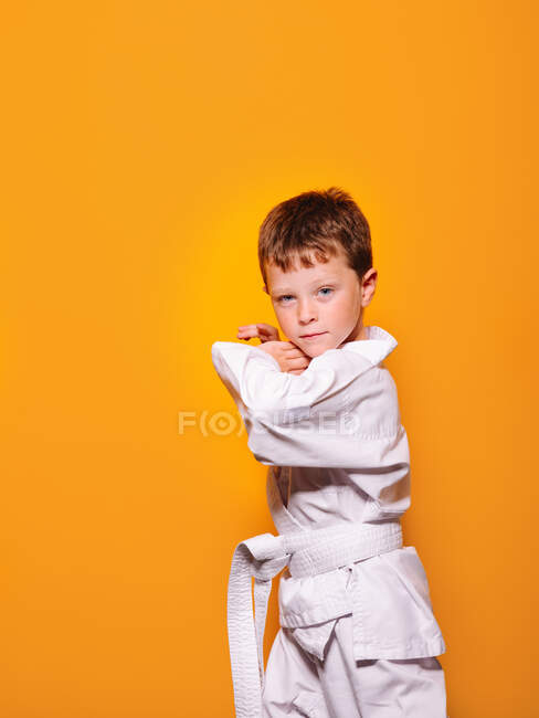 Серйозний хлопчик в білому кімоно робить карате позицію на яскраво-помаранчевому фоні і дивиться на камеру — стокове фото