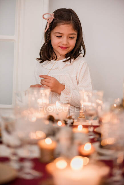 Linda chica en vestido de pie cerca de la mesa festiva y velas relámpago para celebrar las vacaciones de Navidad - foto de stock