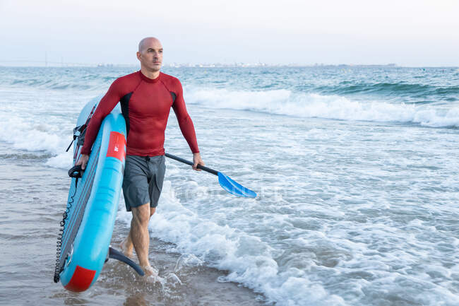 Surfista masculino irreconhecível em fato de mergulho carregando prancha de remo enquanto caminhava olhando para longe na praia — Fotografia de Stock