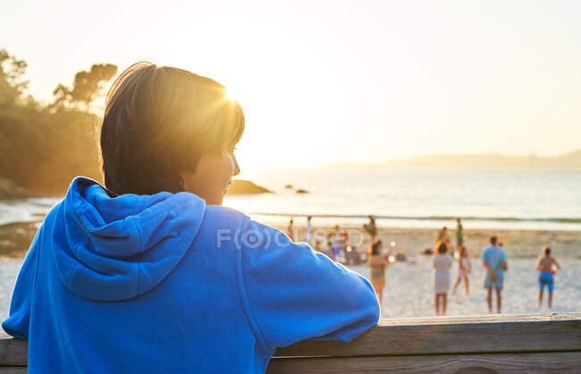 Visão traseira do menino de pé perto de cerca de madeira e admirando o mar cercado por colinas ao pôr-do-sol — Fotografia de Stock