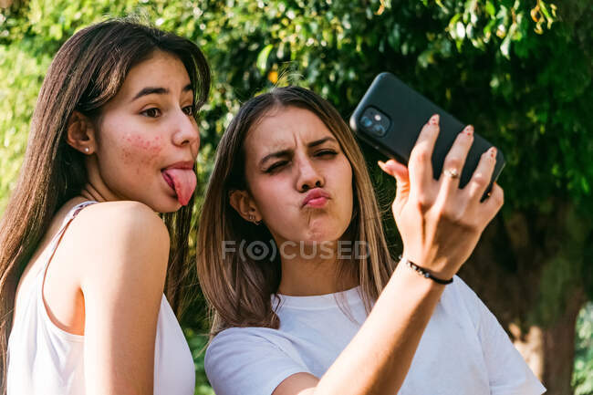Adolescente femenina con labios irritantes cerca de la mejor amiga con acné y lengua fuera tomando autorretrato en el teléfono celular a la luz del sol - foto de stock