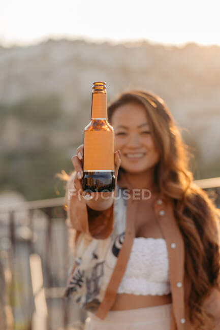 Joven mujer étnica positiva sonriendo brillantemente con botellas de cerveza mientras disfruta de un agradable momento al atardecer en el bar terraza en Capadocia, Turquía - foto de stock