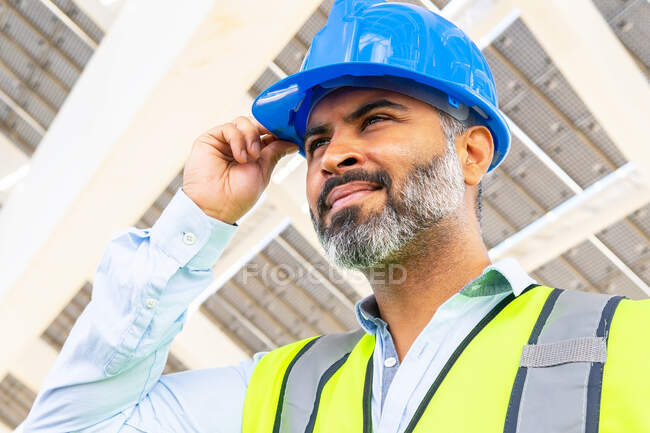 Dal basso di maturo supervisore etnico maschio in giubbotto indossando hardhat mentre guardava lontano in piedi vicino alla centrale solare — Foto stock
