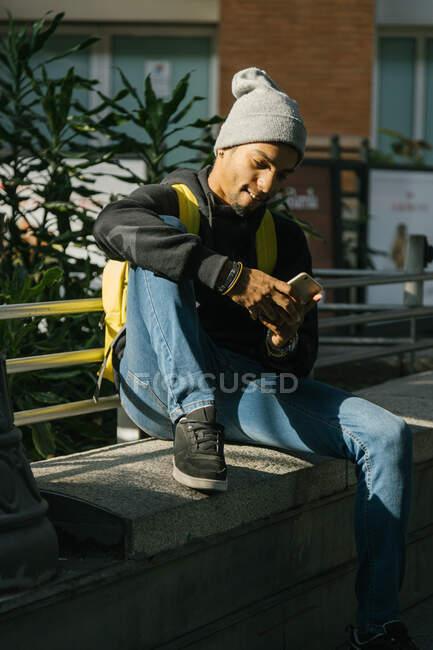 Content African American male hipster with backpack sitting on stone border in street and messaging on mobile phone — Stock Photo