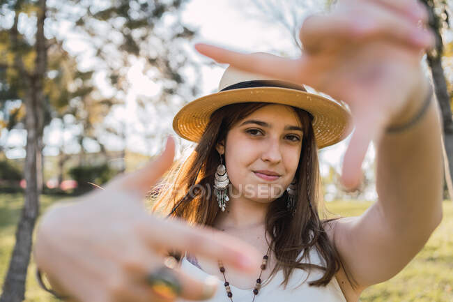 Улыбающаяся девушка-подросток в брекетах демонстрирует жест фотографии, глядя в камеру днем на размытом фоне — стоковое фото