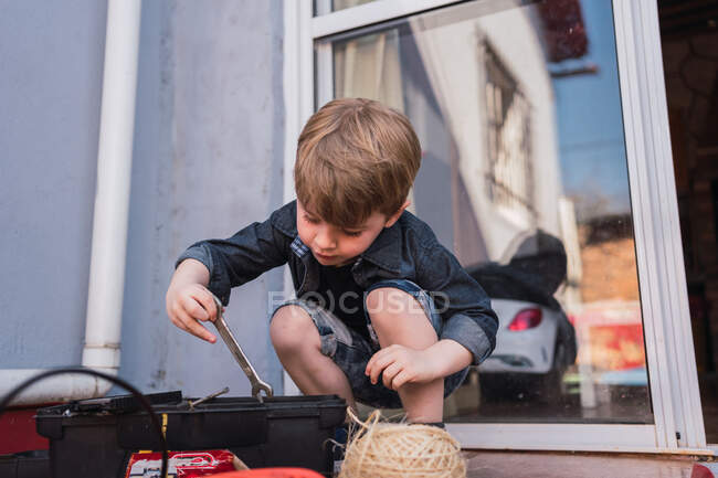 Curious kid taking wrench out of plastic container between glass door and twine ball in daytime — Stock Photo