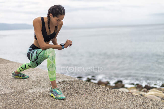 Atleta femenina en forma concentrada en ropa deportiva haciendo ejercicio de embestida hacia adelante mientras calienta los músculos durante el entrenamiento en el paseo marítimo cerca del mar - foto de stock