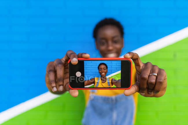 El foco selectivo del teléfono celular en las manos de la mujer alegre afroamericana tomando el autorretrato contra la pared brillante - foto de stock