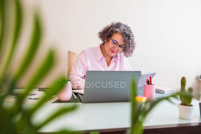 Cheerful elderly female entrepreneur in eyewear with pen speaking on cellphone against netbook on table in workspace — Stock Photo