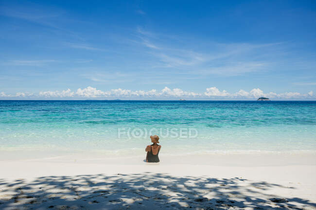 Voltar vista turista feminino em maiô e chapéu de palha sentado em mar transparente durante a viagem na Malásia — Fotografia de Stock