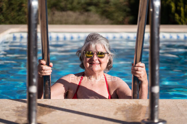 Femme âgée positive en maillots de bain et lunettes de soleil descendant dans la piscine et tenant des mains courantes en acier inoxydable tout en se relaxant dans la journée ensoleillée — Photo de stock