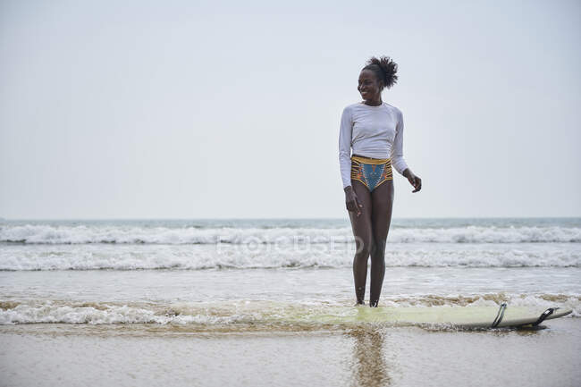 Веселая молодая спортсменка в бикини, стоящая на берегу моря против доски для серфинга, глядя вдаль под легким небом — стоковое фото