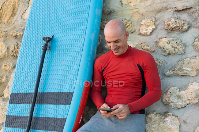 Hombre surfista en traje de neopreno apoyado en la pared de piedra de navegación en el teléfono inteligente con paleta y tabla SUP mientras se prepara para navegar en la orilla del mar - foto de stock