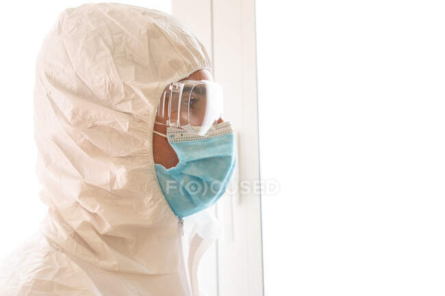 Adult male medic in personal protective equipment with glasses and sterile mask looking forward against window in hospital — Stock Photo