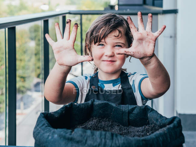 Positive little girl in apron standing at table with flower pot and demonstrating dirty hands on balcony in daytime — Stock Photo