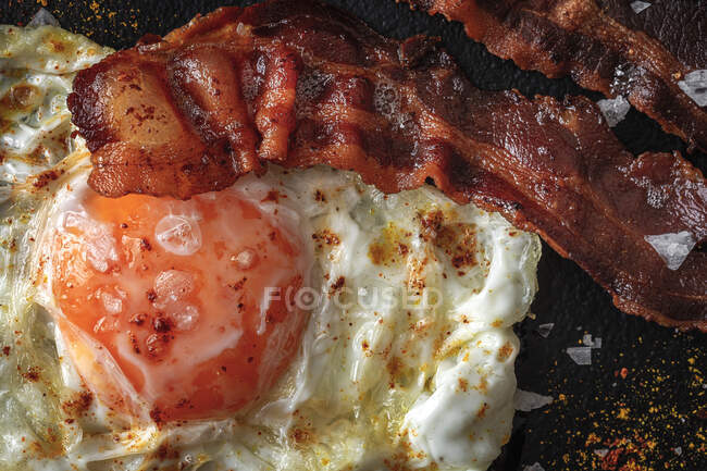 De cima do lado ensolarado acima do ovo com fatias de bacon fritas e condimentos na bandeja escura — Fotografia de Stock