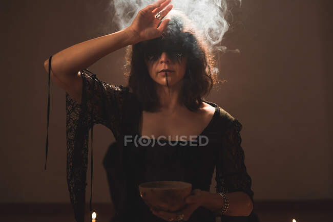 Witch in black dress and with painted face standing with bowl in room with steam during spiritual ritual — Stock Photo