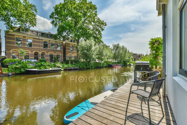 Terraço de madeira do edifício residencial perto do canal do rio localizado na área do subúrbio com árvores verdes no dia ensolarado — Fotografia de Stock