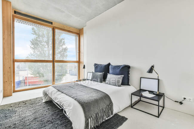 Kreative Gestaltung des Schlafzimmers mit Kissen auf Bettdecke auf dem Bett zwischen Tisch mit Lampe und Fenster im Haus — Stockfoto