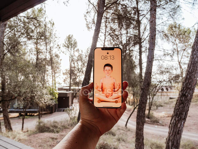 Cultivo irreconocible persona sosteniendo el teléfono móvil con la imagen de un niño haciendo yoga en la pantalla - foto de stock