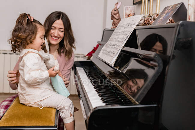 Alegre mamá interactuando con encantadora chica sosteniendo el juguete contra el piano con la hoja de música en casa - foto de stock