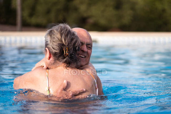 Hombre calvo sonriente abrazando a una mujer sin camisa mientras nadan juntos en agua limpia de la piscina - foto de stock