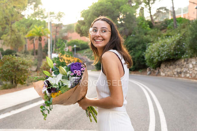 Vista laterale di amichevole giovane femmina in occhiali con bouquet floreale in fiore guardando lontano sulla strada della città — Foto stock