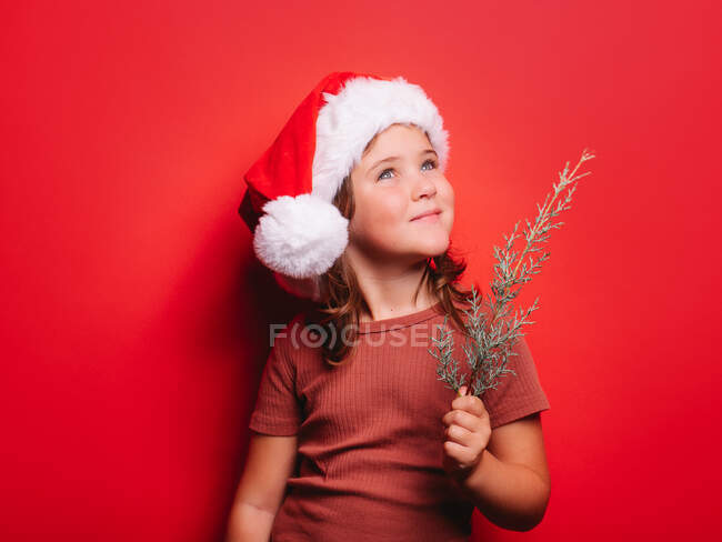 Petite fille mignonne en vêtements décontractés et chapeau de Père Noël tenant rameau de sapin et regardant loin sur fond rouge — Photo de stock