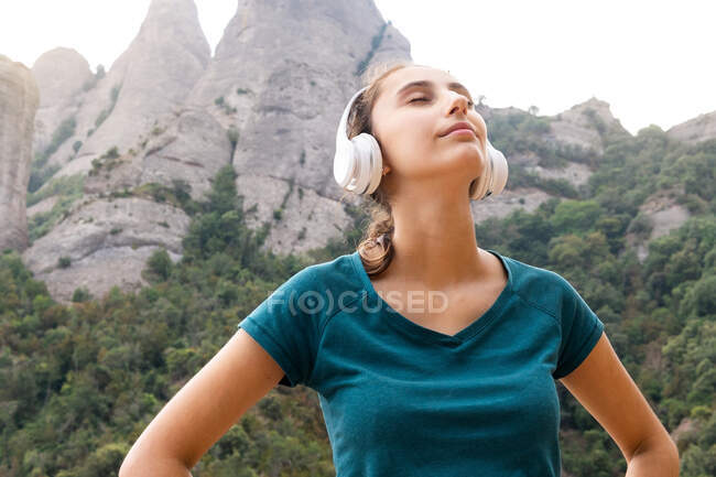 Dreamy young female tourist with closed eyes enjoying song from wireless headset against Montserrat and trees in Spain — Stock Photo