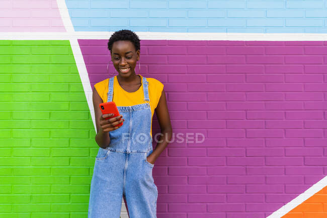 Afro-americano fêmea em macacão jeans em pé perto de parede colorida e celular de navegação — Fotografia de Stock