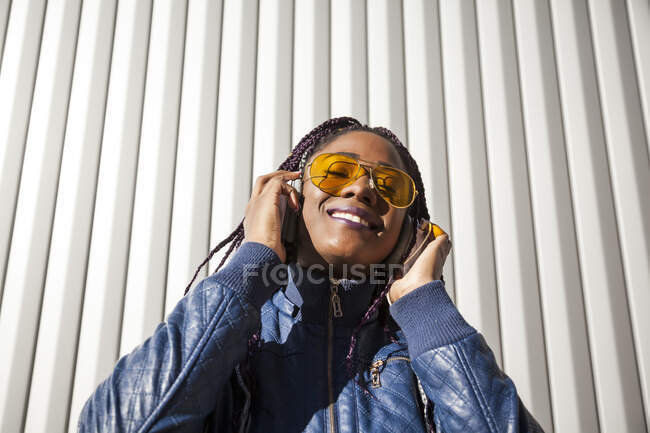 Happy young African American female with Afro braids dressed in blue jacket and stylish sunglasses enjoying music through earphones while chilling in sunlight against striped wall — Stock Photo